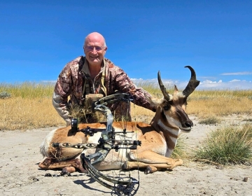 A bowhunter, dressed in camouflage and gear, poses with a pronghorn antelope and his compound bow, showing the rewards of an SNS Outfitter & Guides archery hunt.